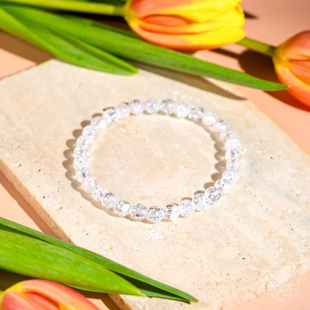 Ice Quartz bracelet with 6mm clear, faceted quartz beads reflecting light, arranged in a circle on a beige stone tile with orange tulips nearby