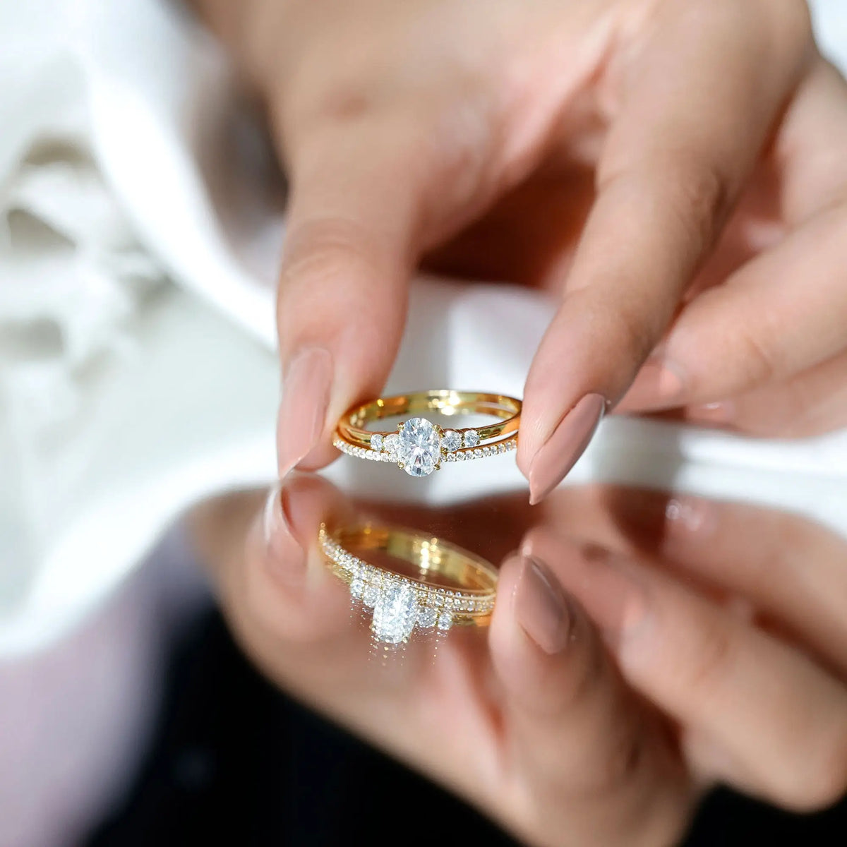 Lab Grown Diamond gold engagement ring with oval center stone and round side diamonds held by hand reflecting on glossy surface